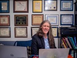 A man sits at his desk smiling with many diplomas on the wall behind him.