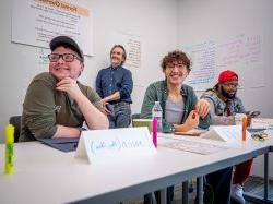 Three people sit at a table and one leans against a wall as they listen to a workshop presentation.